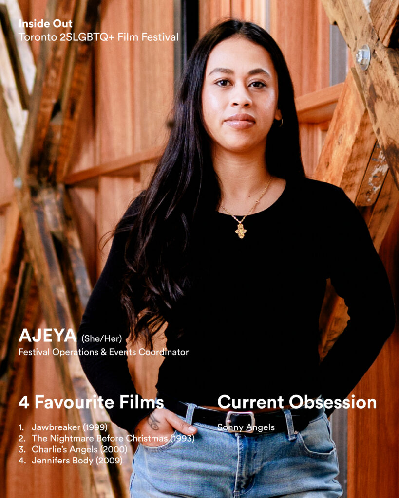 A person with long dark hair wearing a black top and jeans stands confidently in a wooden structure.