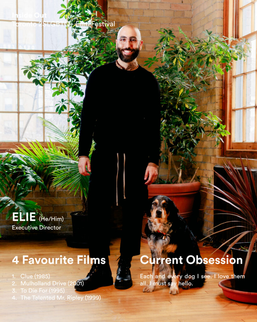 A smiling man with a beard, dressed in black, stands indoors beside a black and brown dog. Sunlight streams through large windows onto exposed brick.
