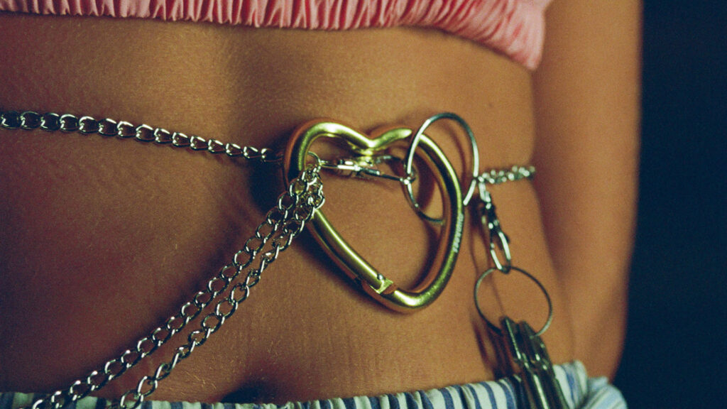 Close-up of a person’s midriff wearing a metal chain belt with a heart-shaped clasp, layered over blue and white striped bottoms and a pink ruched top.