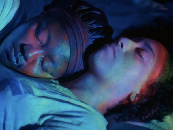 Two people lie close together, sleeping peacefully. One rests their head on the other's chest. Soft blue and purple lighting creates a calm, intimate atmosphere.