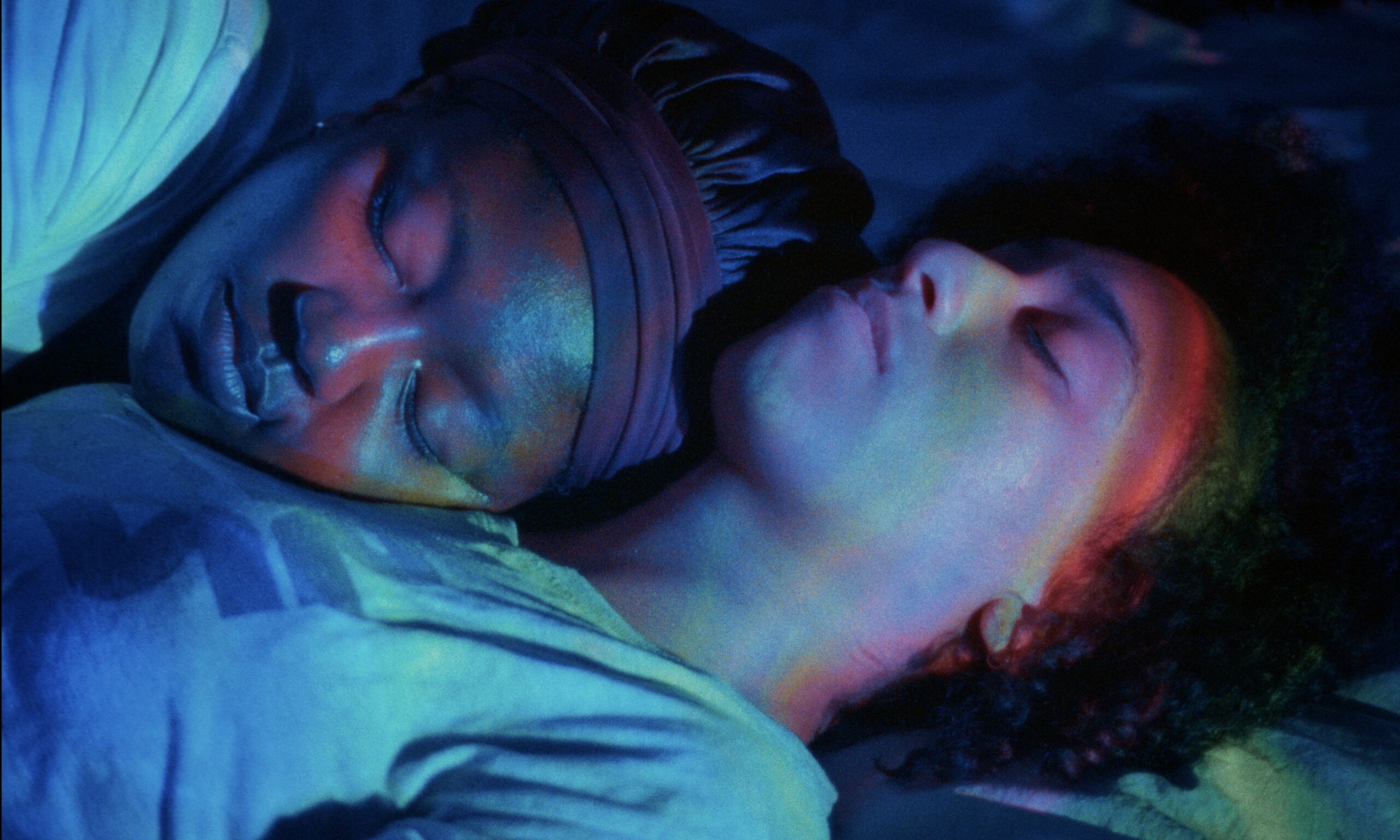 Two people lie close together, sleeping peacefully. One rests their head on the other's chest. Soft blue and purple lighting creates a calm, intimate atmosphere.