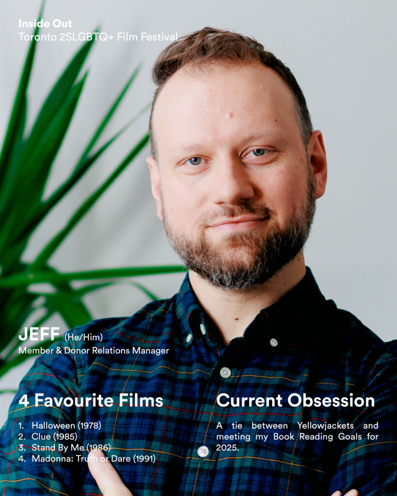 A bearded person with light skin and short hair stands in front of a leafy green plant, wearing a blue checked shirt.
