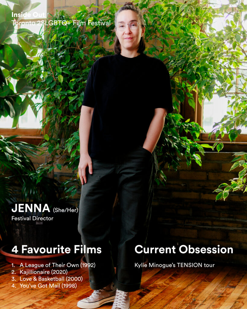 A person with glasses and long hair, wearing a black t-shirt and dark pants, stands indoors by plants and brick walls.