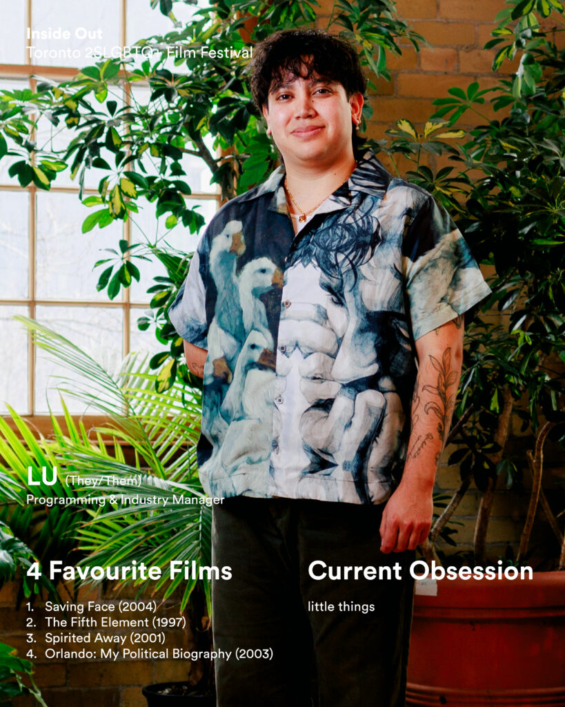 A person with short dark hair and tattoos stands in front of plants, wearing a shirt with a swan print.