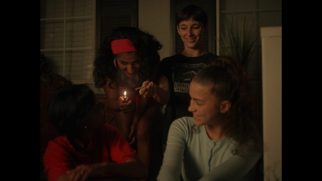 Four teens gather outside at night; one holds a lighter with a small flame, while the others smile and laugh, creating a warm, friendly, and playful atmosphere.