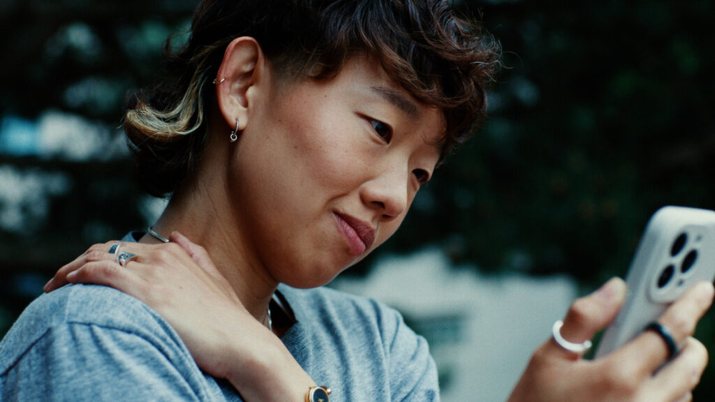 A person with short, wavy hair and earrings smiles gently while looking at a smartphone. They wear a gray shirt and rings, with one hand resting on their shoulder. The background is blurred greenery.