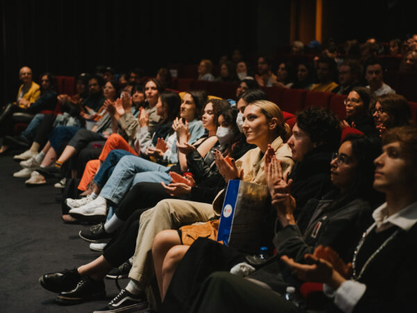 A diverse audience sits in rows of theater seats, attentively watching and applauding an unseen event or presentation on stage. The lighting is dim, focusing on the spectators’ engaged expressions.