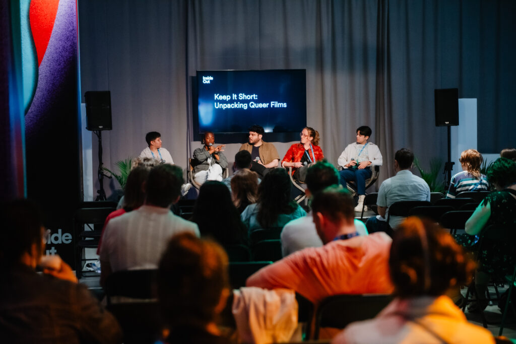 A panel of five people sits on stage discussing in front of an audience. Behind them, a screen displays the text, “Keep It Short: Unpacking Queer Films.” The room is dimly lit with a casual, attentive atmosphere.