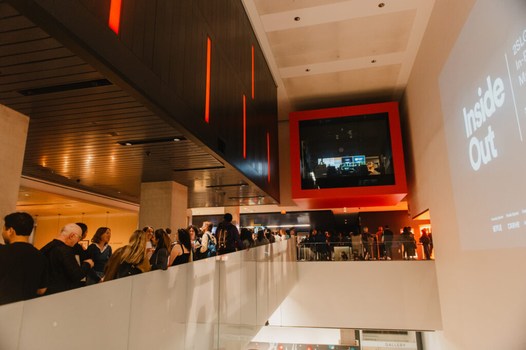 A crowd of people stand in line along an indoor balcony at the TIFF Lightbox venue. On the wall, a large projected sign reads "Inside Out," with people visible on an upper level socializing and mingling in line for the film.