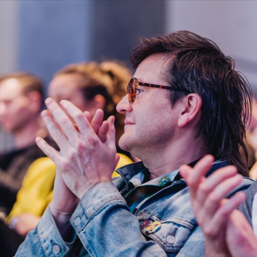 A group of people sitting and clapping, with a person in the foreground wearing sunglasses and a denim jacket, focused on an event or presentation.