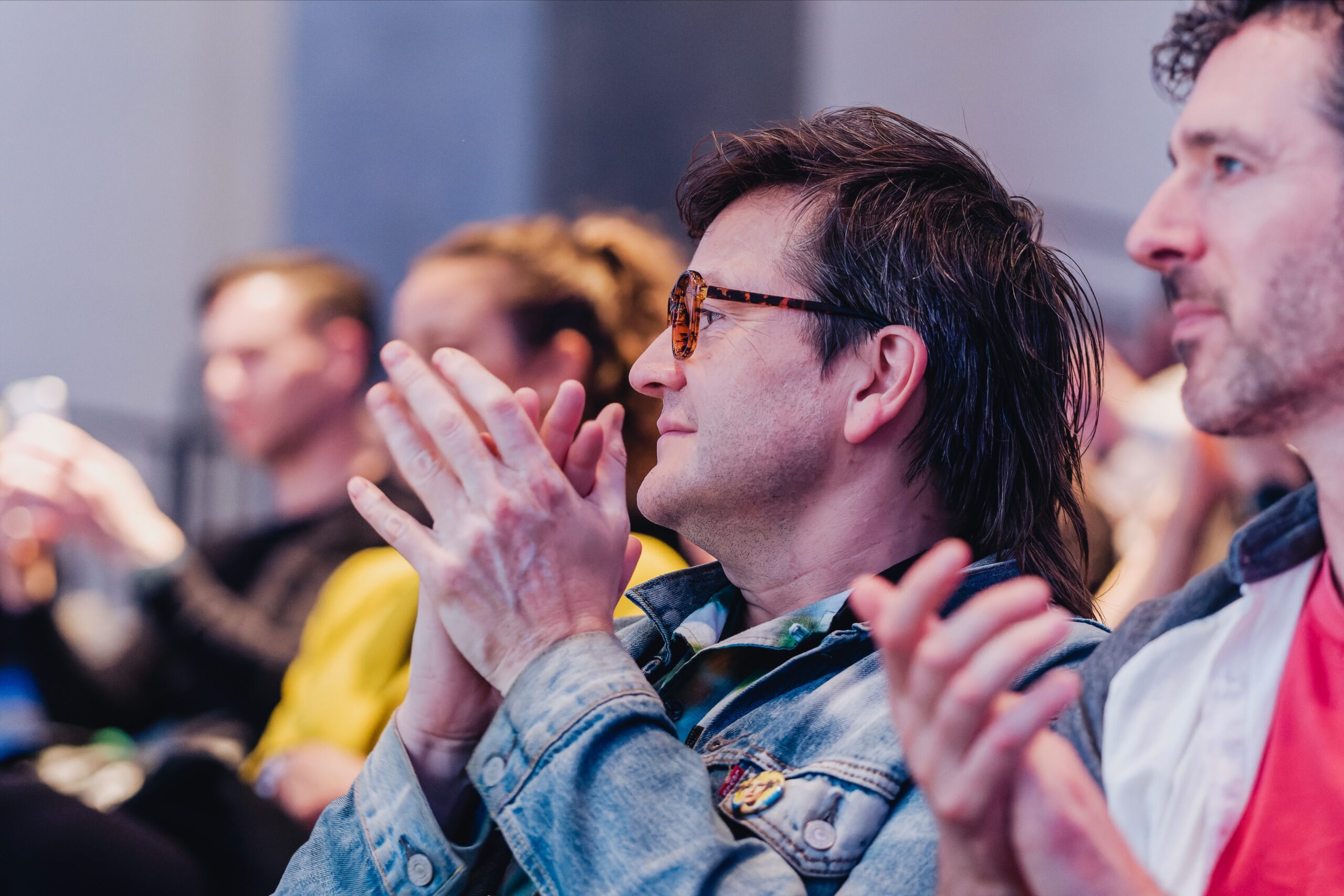A group of people sitting and clapping, with a person in the foreground wearing sunglasses and a denim jacket, focused on an event or presentation.
