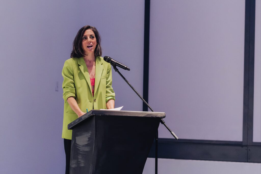 A woman in a bright green blazer speaks at a podium with a microphone, holding papers, against a plain light-colored background.