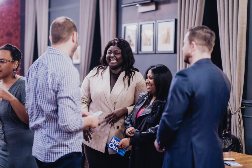 A group of four adults stand indoors, engaged in conversation and smiling. One person holds a blue booklet that says "Inside Out." Framed pictures and draped curtains are visible in the background.