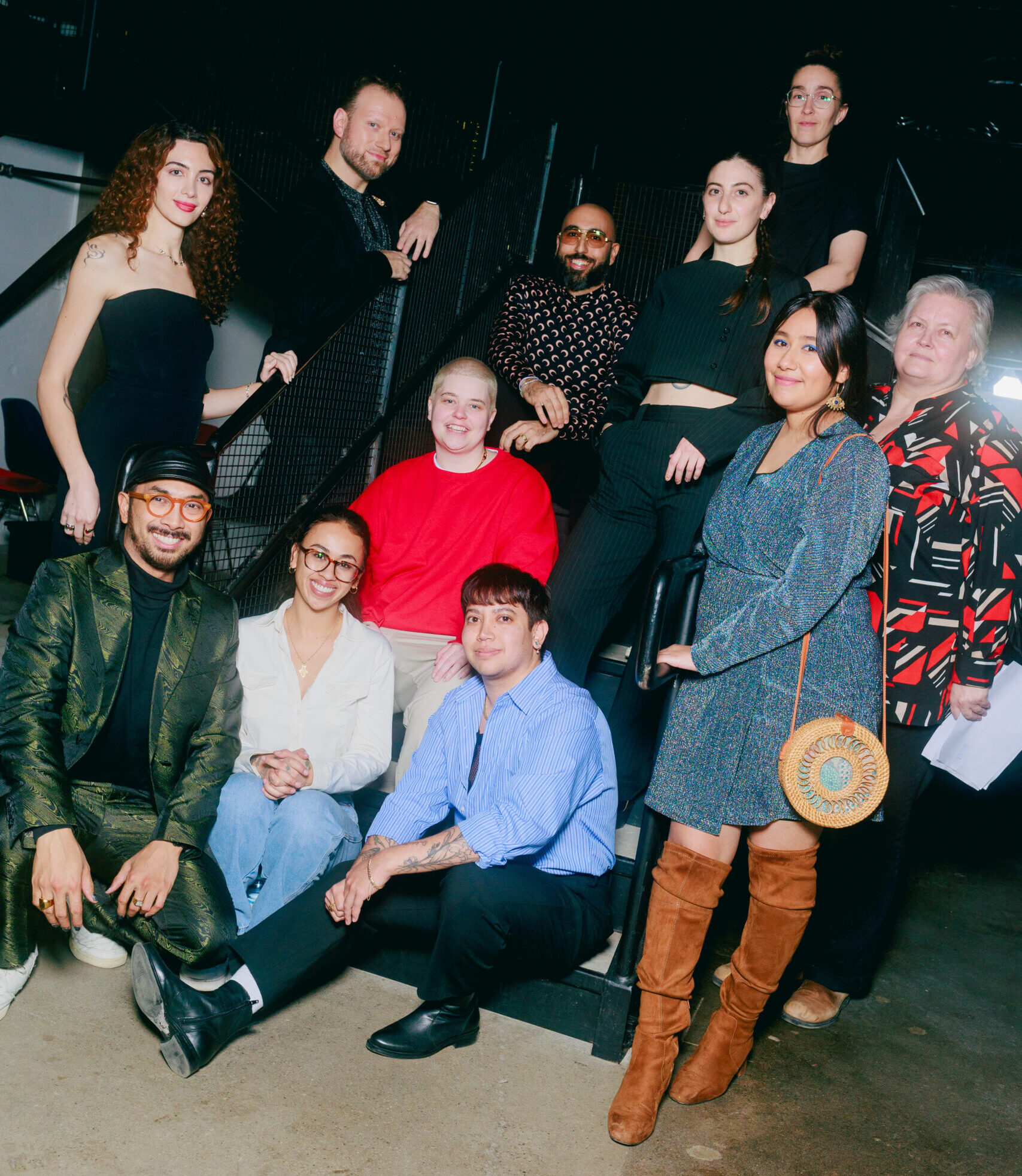 A group of eleven people pose together on and around a staircase indoors, smiling at the camera. The lighting is bright, and everyone is dressed in stylish, casual to semi-formal attire.