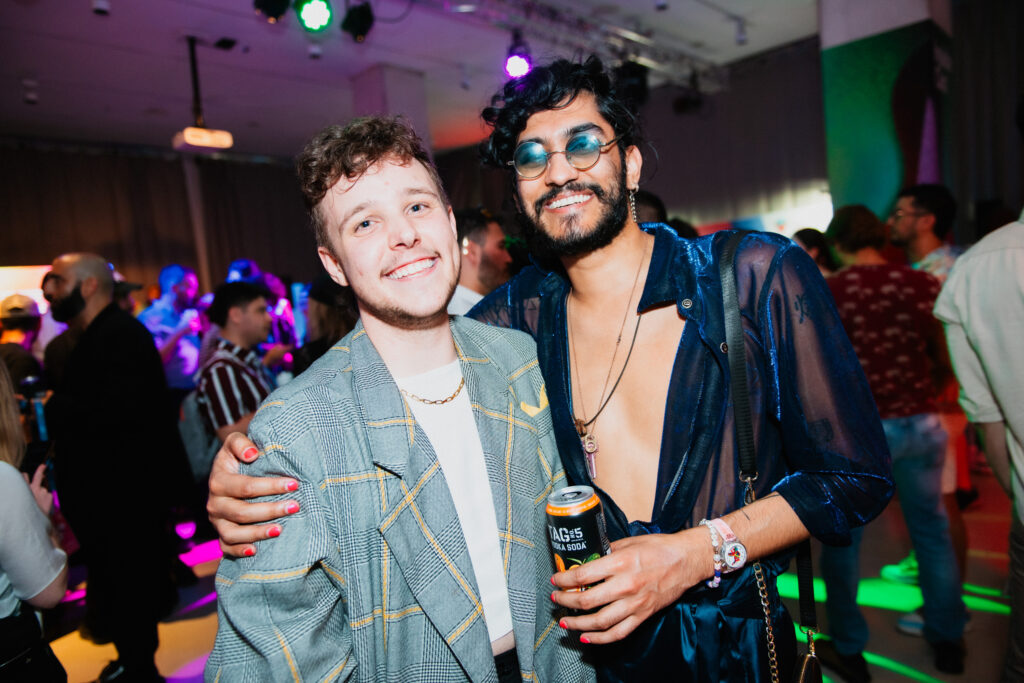 Two people stand close together at a lively party, smiling at the camera. One holds a drink can, both wear stylish, bold outfits. Colorful lights and other guests fill the vibrant background.