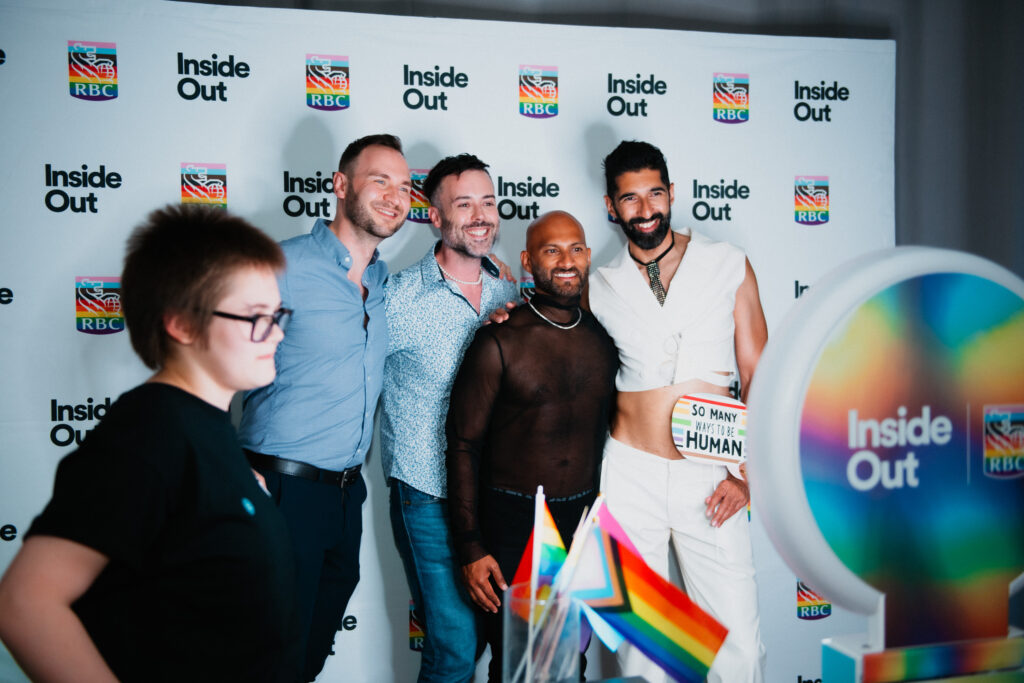 Four people pose and smile in front of an Inside Out RBC backdrop at an event, with rainbow pride flags visible in the foreground and a person with short hair standing to the left.