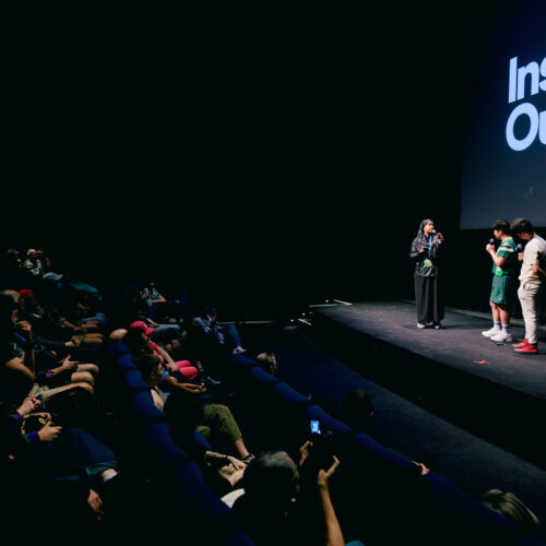 Four people stand on stage under a screen reading “Inside Out” as an audience watches in a dark theater. The room is dimly lit, and one person is interviewing the others.