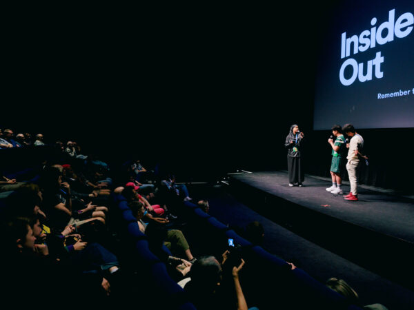 Four people stand on stage under a screen reading “Inside Out” as an audience watches in a dark theater. The room is dimly lit, and one person is interviewing the others.
