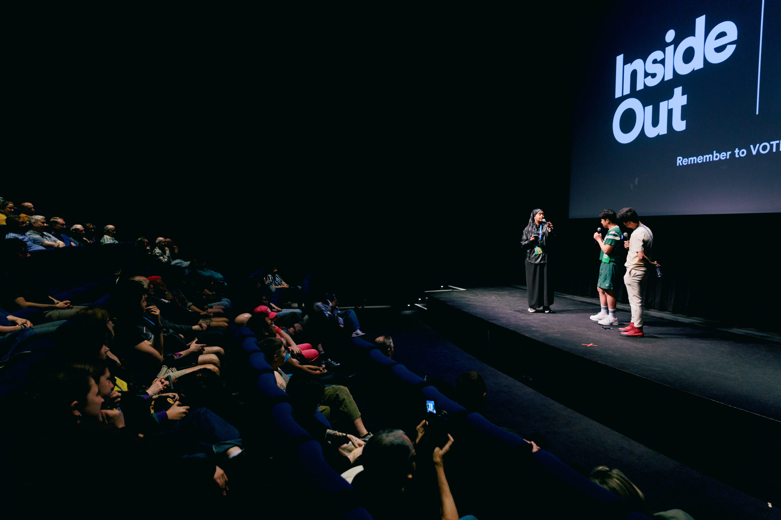 Four people stand on stage under a screen reading “Inside Out” as an audience watches in a dark theater. The room is dimly lit, and one person is interviewing the others.