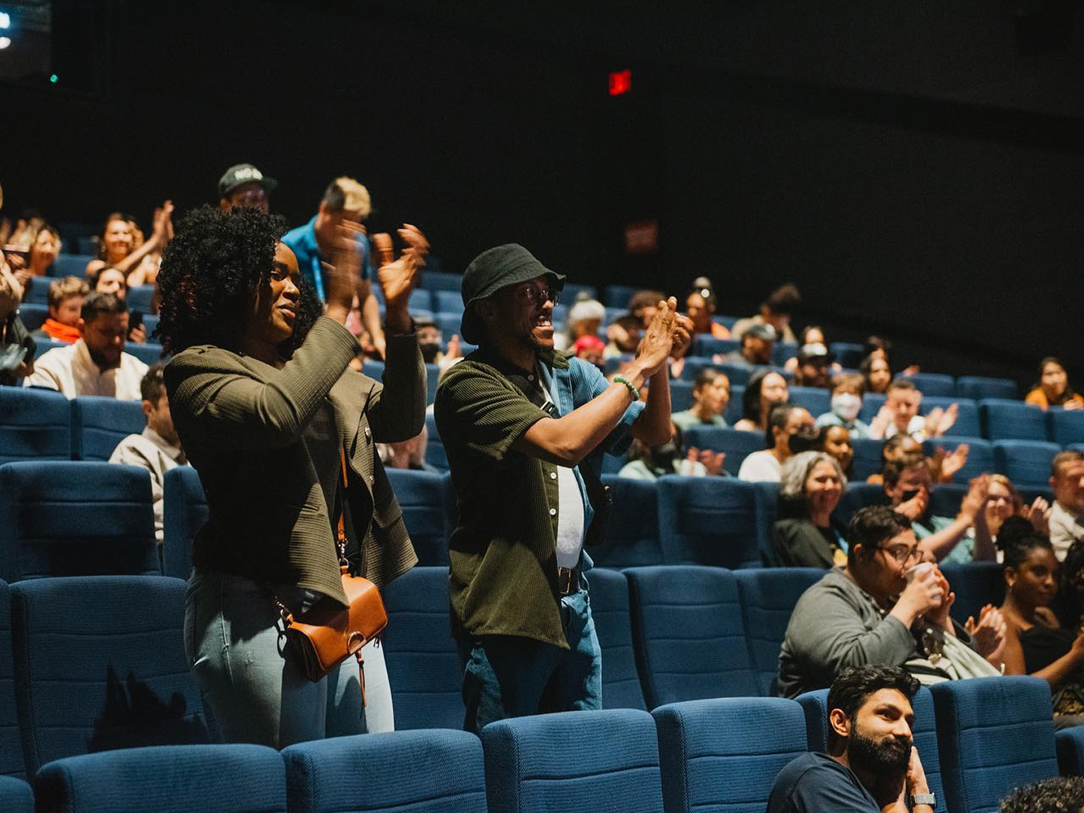 A crowd in cinema seats stand and applaud towards the stage. Cinem seats are blue.