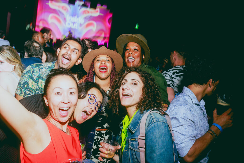 A group of six people smiling and posing for a selfie at a lively party, holding drinks, with colorful lights and a crowd in the background.