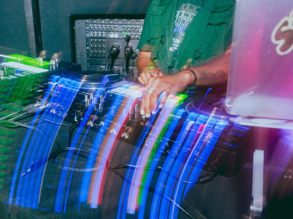 A DJ stands behind turntables and a mixer, with colorful light trails from their moving hands creating a dynamic, energetic effect. The DJ's face is not visible; only their torso and arms are shown.