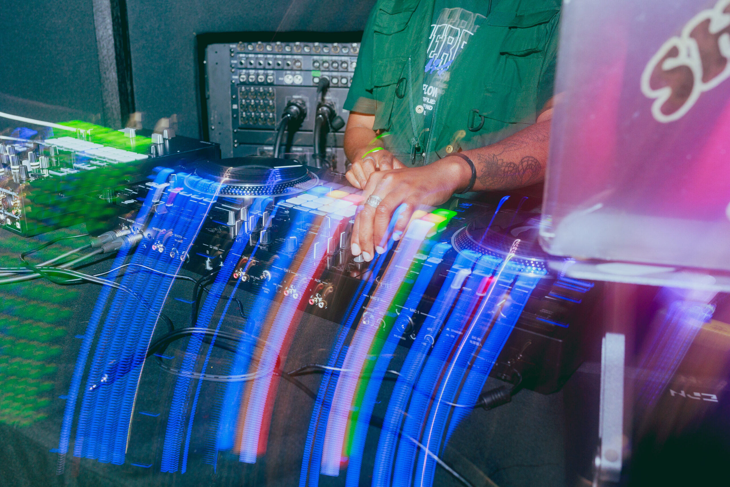 A DJ stands behind turntables and a mixer, with colorful light trails from their moving hands creating a dynamic, energetic effect. The DJ's face is not visible; only their torso and arms are shown.