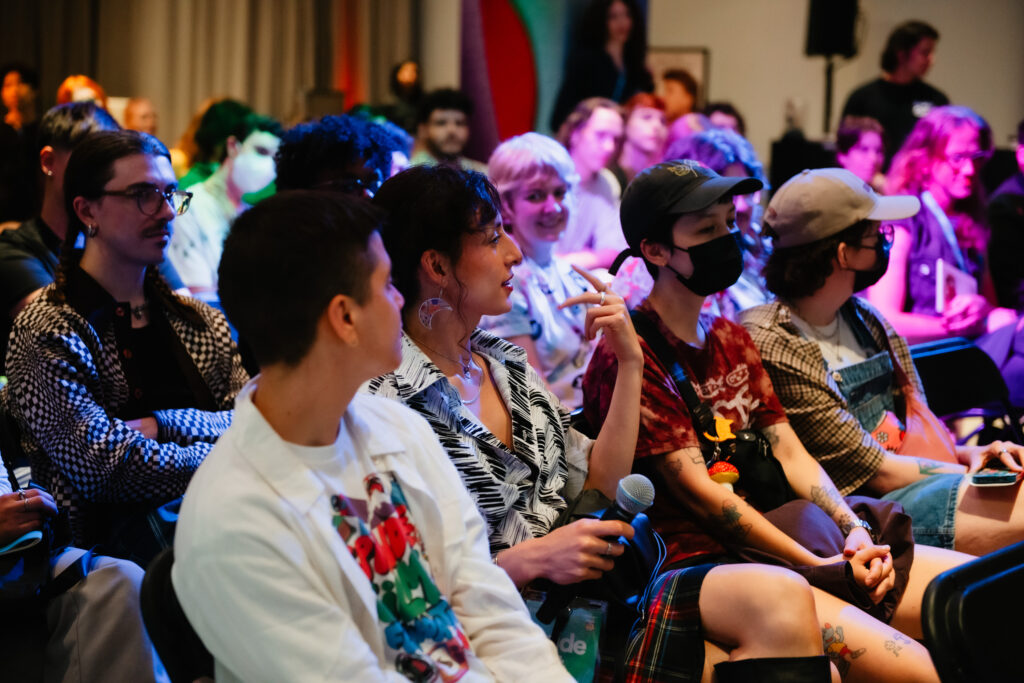 A group of people sit closely together in an indoor event. One person in the middle holds a microphone and appears to be speaking, while others listen attentively. The mood is vibrant and casual, with colorful lighting.