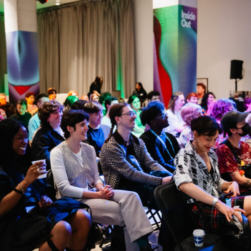 A diverse group of people sit closely together in rows at an indoor event, smiling and engaged. Colorful lighting and abstract decor create a lively atmosphere. Some attendees hold drinks, and a curtain hangs in the background.