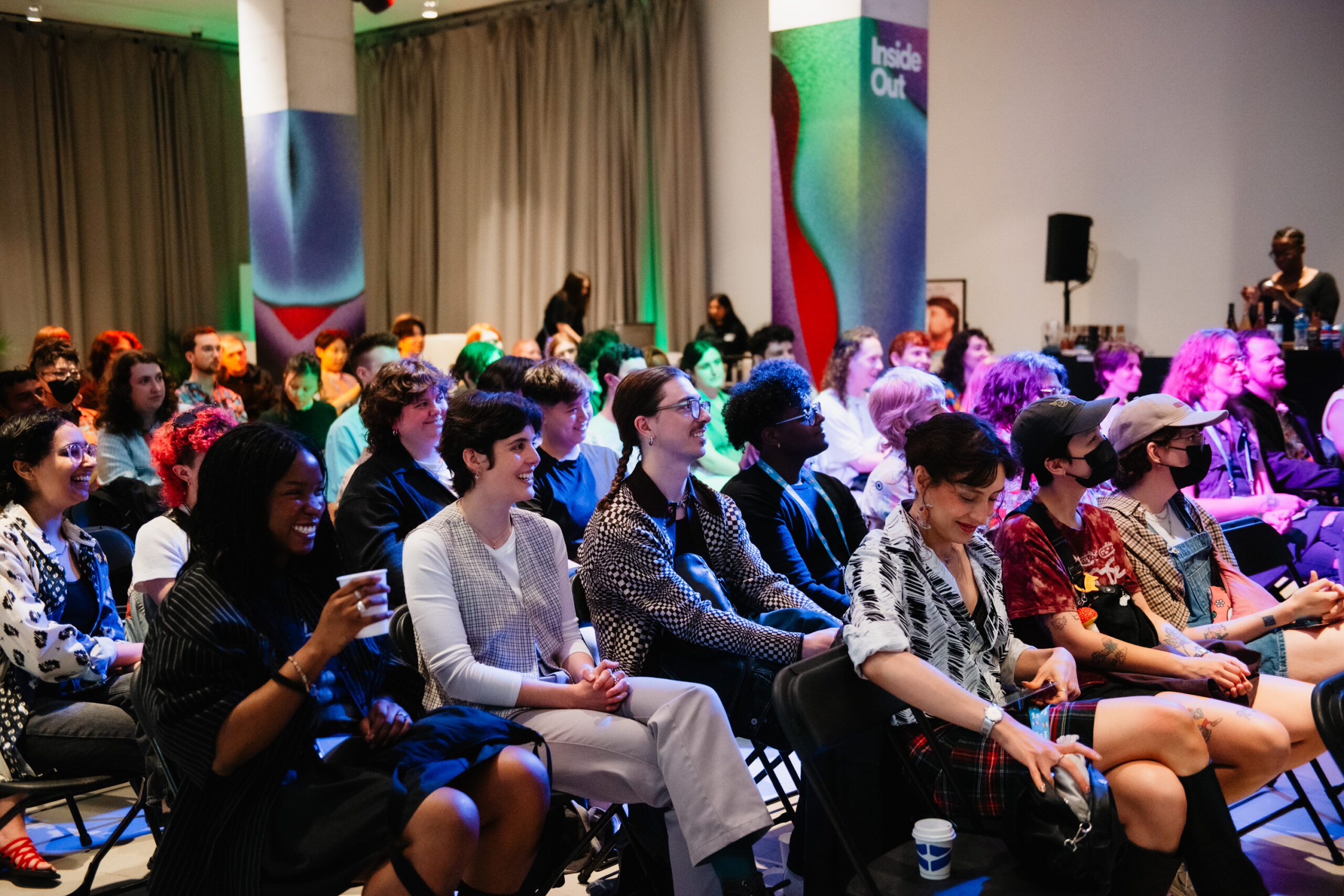 A diverse group of people sit closely together in rows at an indoor event, smiling and engaged. Colorful lighting and abstract decor create a lively atmosphere. Some attendees hold drinks, and a curtain hangs in the background.