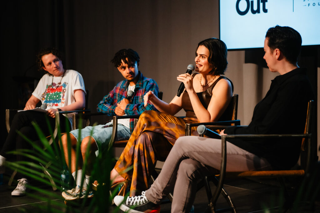 Four people sit on stage in chairs having a panel discussion. One person, holding a microphone, is speaking while the others listen attentively. A plant is partially visible in the foreground.