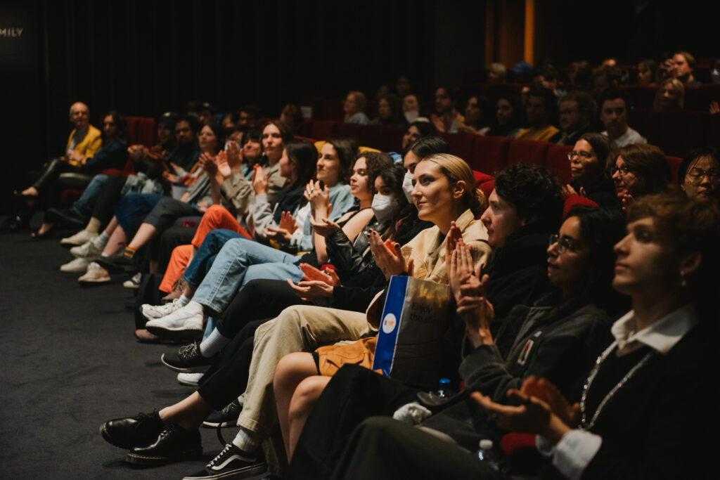 A diverse audience sits in rows of red theater seats, attentively watching and applauding something off-camera. The dim lighting highlights their engaged expressions and casual attire.