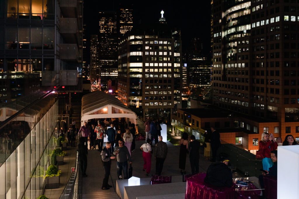 A rooftop party at night with guests mingling under a white tent, surrounded by tall city buildings illuminated by lights. Some people stand near the edge, enjoying the view of the city skyline in the background.