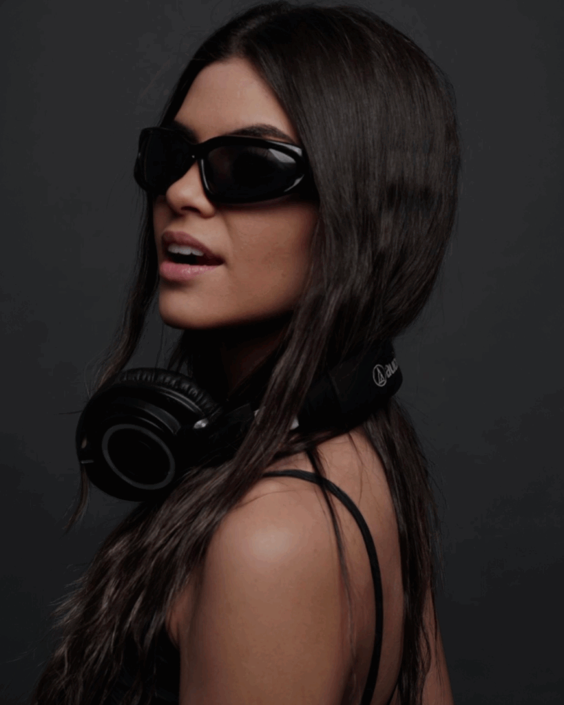 A woman with long dark hair wears black sunglasses and headphones around her neck. She is dressed in a black strappy top and poses against a dark background.
