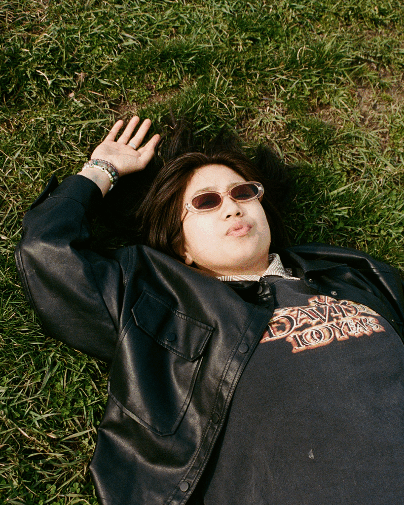 A person with long hair wearing sunglasses, a black leather jacket, and a graphic t-shirt lies on grass with one hand raised and the other by their side.