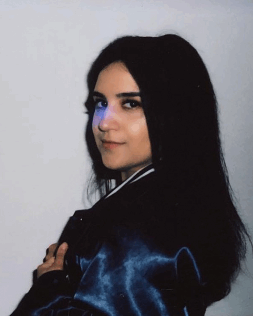 A woman with long dark hair stands sideways against a plain light background, gazing at the camera and wearing a dark jacket with her hand holding the collar.