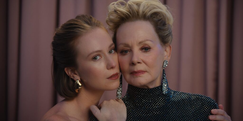 Two women pose closely together in front of pink curtains. One is older, wearing a sparkly black top and long earrings, while the younger woman leans on her shoulder, both looking calmly at the camera.
