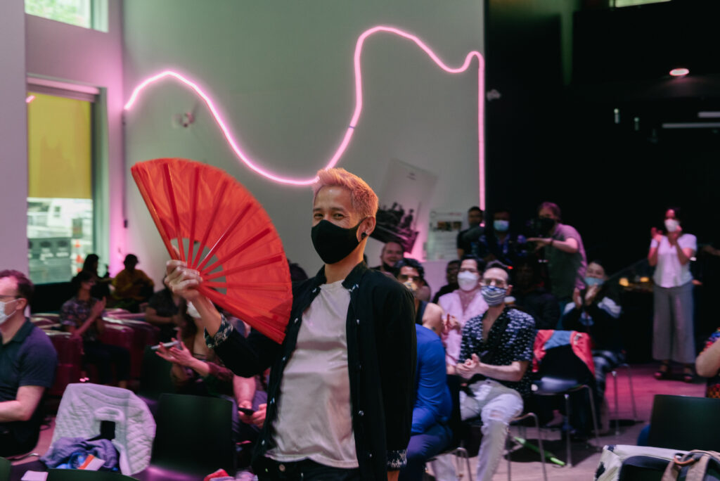 Person with orange hair and a black face mask stands indoors, holding a large red fan, while an audience, many also masked, watches. Neon pink lights decorate the wall behind them.