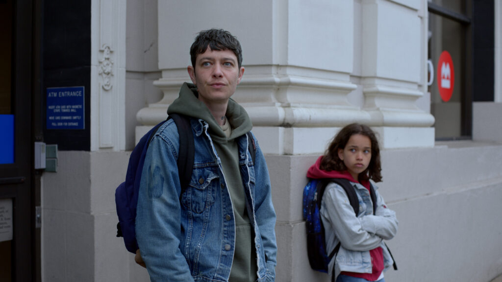 An adult wearing a denim jacket and backpack stands next to a child with curly hair and a backpack, both leaning against a building with white columns. The adult looks to the side while the child gazes ahead.