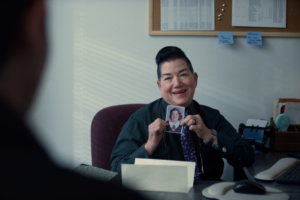 A person sitting at an office desk smiles and holds up a photo of someone in a suit and tie. The desk has office supplies, and a corkboard with papers and sticky notes is on the wall behind them.