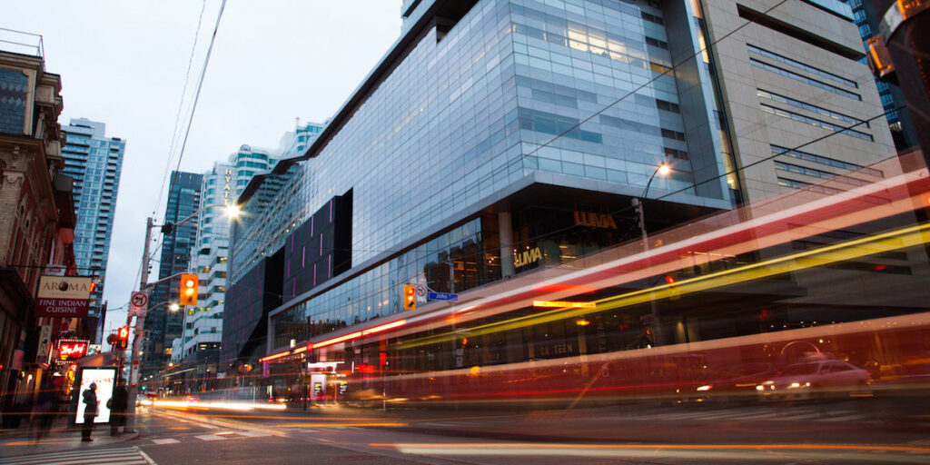 A busy city street with blurred streaks of light from passing streetcars, modern glass buildings, and tall skyscrapers in the background. Pedestrians wait at the crosswalk as evening approaches.