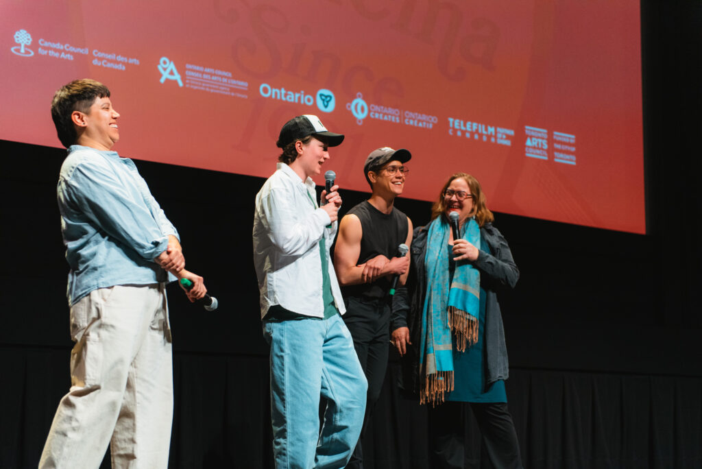 Four people stand onstage with microphones, smiling and laughing, in front of a large red screen displaying logos and text, suggesting a film or arts event.