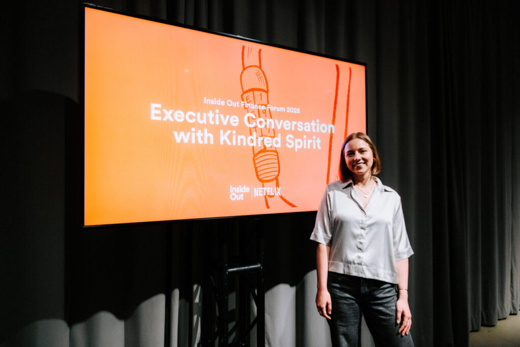 A woman in a light blouse stands smiling next to a large screen displaying "Executive Conversation with Kindred Spirit, Inside Out Film Festival, Netflix," against an orange background.