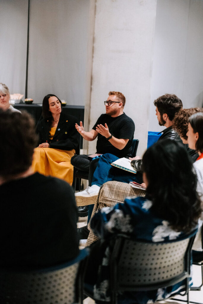 A group of people sits in a circle having a discussion. One person in the center gestures with his hands while speaking, and others listen attentively. The setting appears to be an informal meeting or workshop.