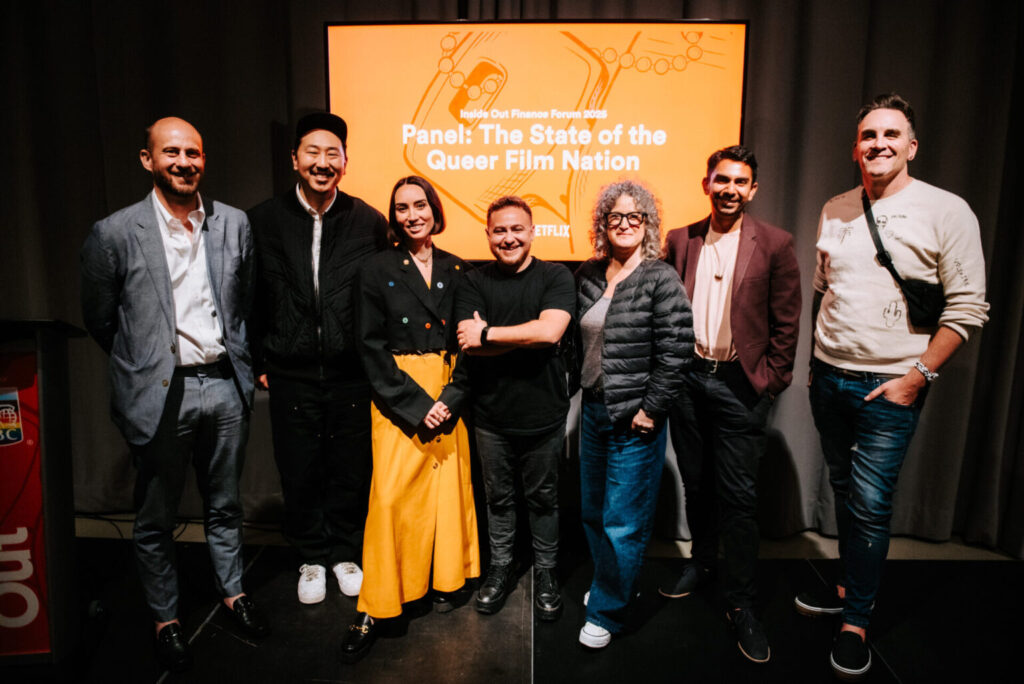 Seven people stand together smiling in front of a screen that reads “Panel: The State of the Queer Film Nation.” The group is casually dressed, and the setting appears to be an indoor film or discussion event.