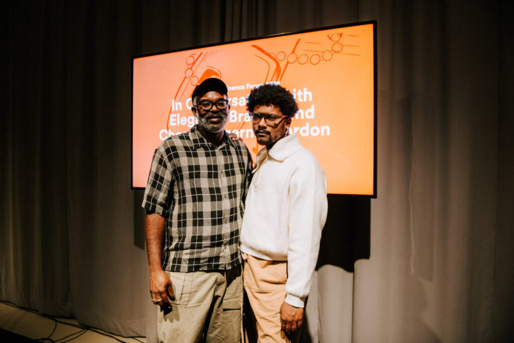 Two men stand side by side in front of a screen with orange background and white text. One wears a plaid shirt and beanie, the other wears glasses and a white shirt. Both face the camera and appear relaxed.