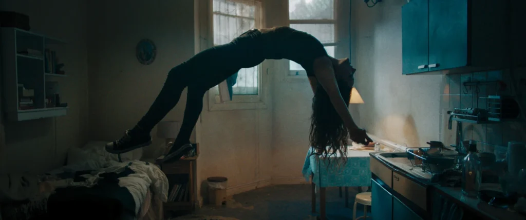 A person with long hair floats horizontally in midair above a small, dimly lit room with a bed, table, and kitchen area, creating a surreal, mysterious atmosphere.