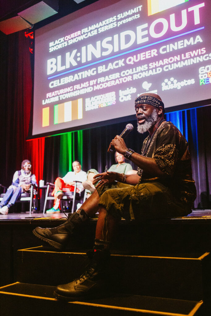 A speaker sits on the edge of a stage, holding a microphone, with a panel seated behind him. A colorful banner above reads “BLK: INSIDE OUT: Celebrating Black Queer Cinema.” The stage is lit with red, green, and blue lights.
