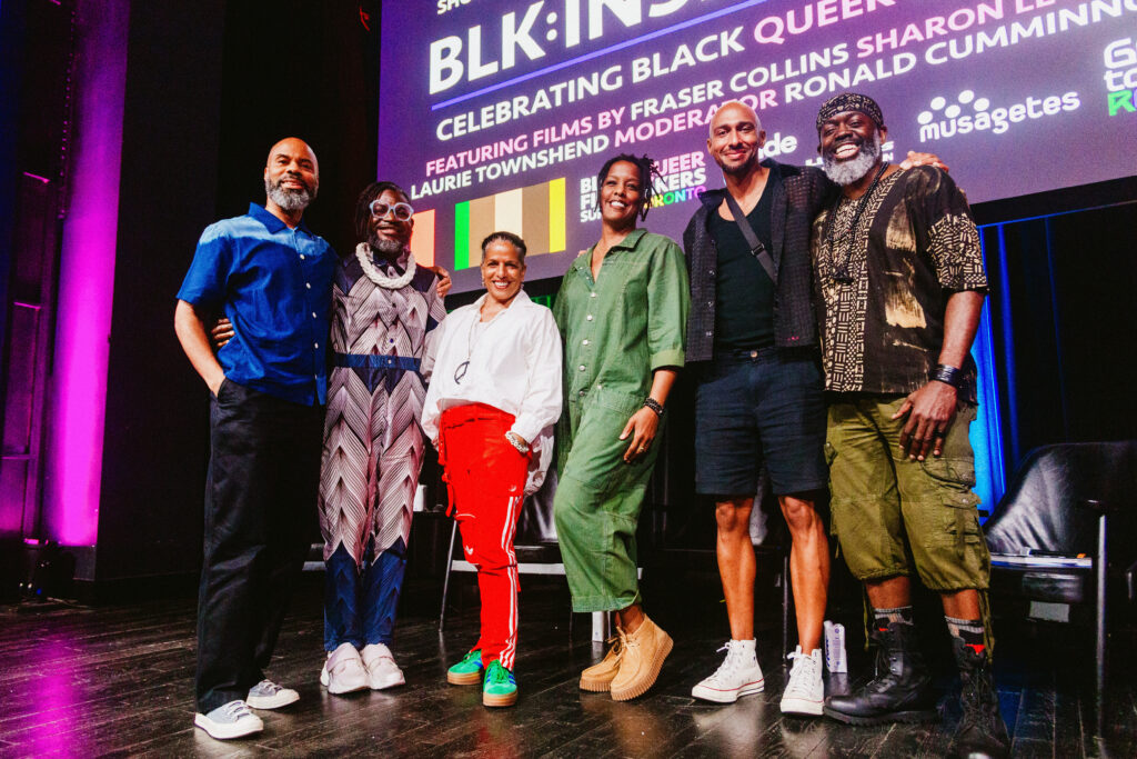 Six people stand together smiling on a stage in front of a colorful backdrop reading "BLK: INSIGHT Celebrating Black Queer." They are dressed in vibrant, stylish outfits, and appear relaxed and happy.