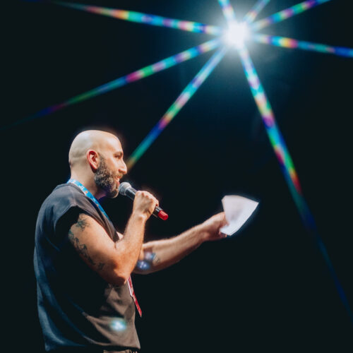 A person with a beard and tattoos speaks into a microphone on stage, holding papers. Bright stage lights create a starburst effect above them against a dark background.